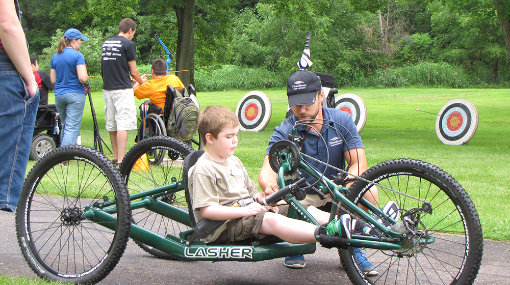 Event staff help child with a hand-pedal tricycle