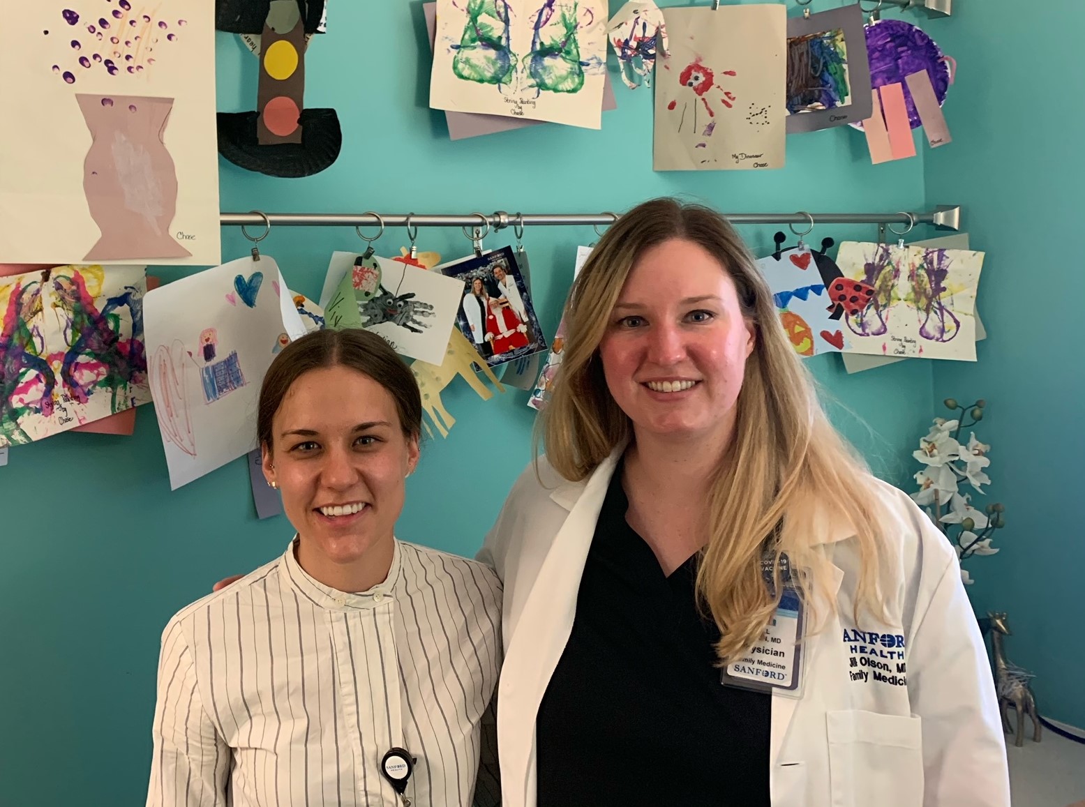Two women smile in front of a wall adorned with children's artwork.