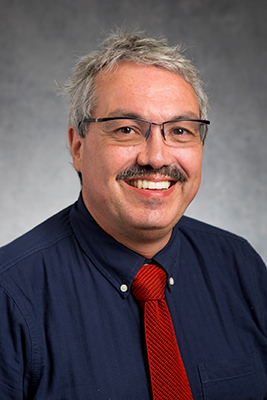 Dr. Paul bigliardi wearing a blue button up shirt and red tie. He has grey short hair, glasses, and a mustache