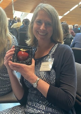 photo of Dr. Christina Boull, medium blonde hair, smiling, and wearing a blue dress with a cardigan, and holding an apple award 