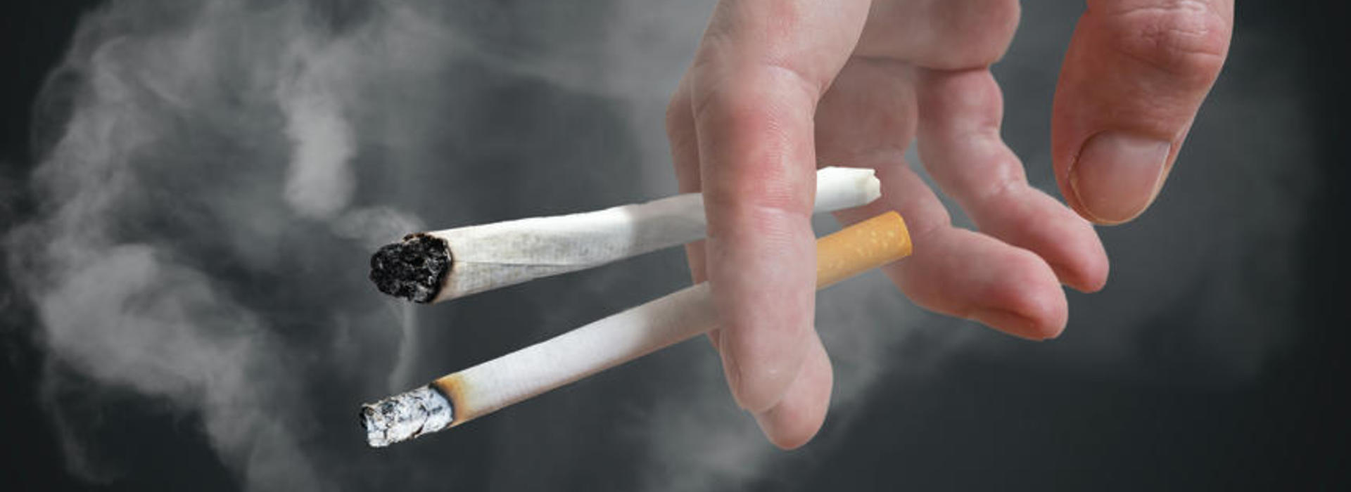 Man (smoker) is holding cigarette in hand on black background. Smoke around.