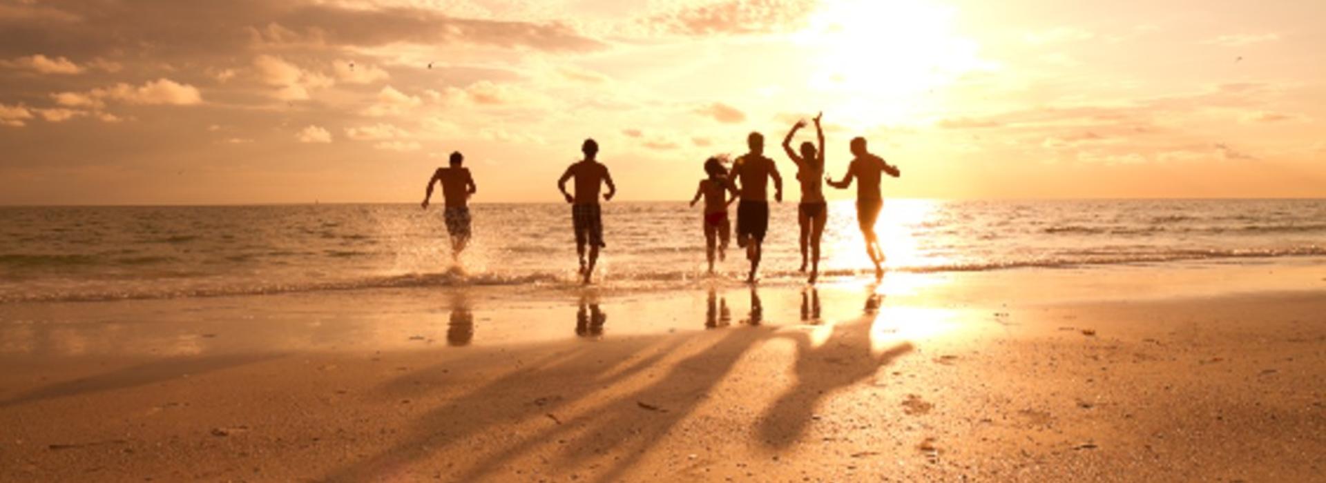 A shot of a group of people in swimwear running toward water on a beach while the sun sets in the background.
