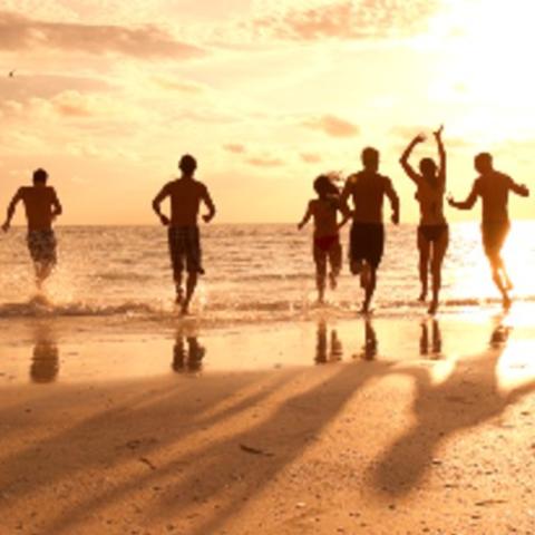 A shot of a group of people in swimwear running toward water on a beach while the sun sets in the background.